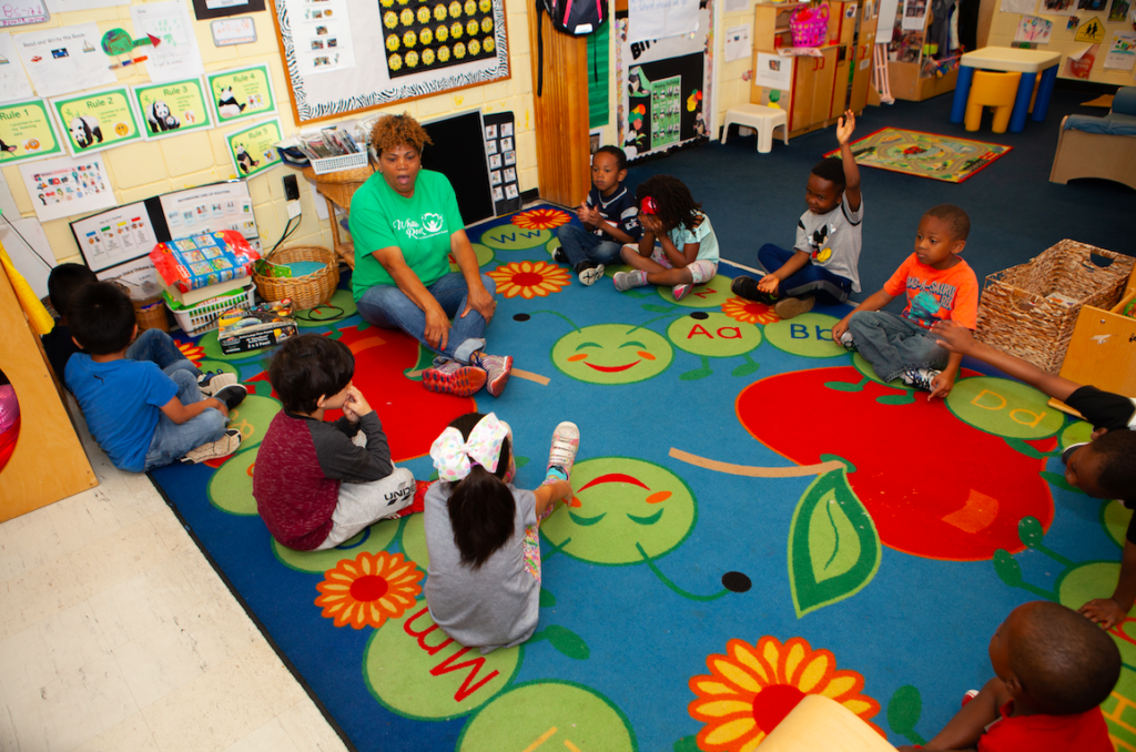 A teacher and several students sit in a circle on a rug