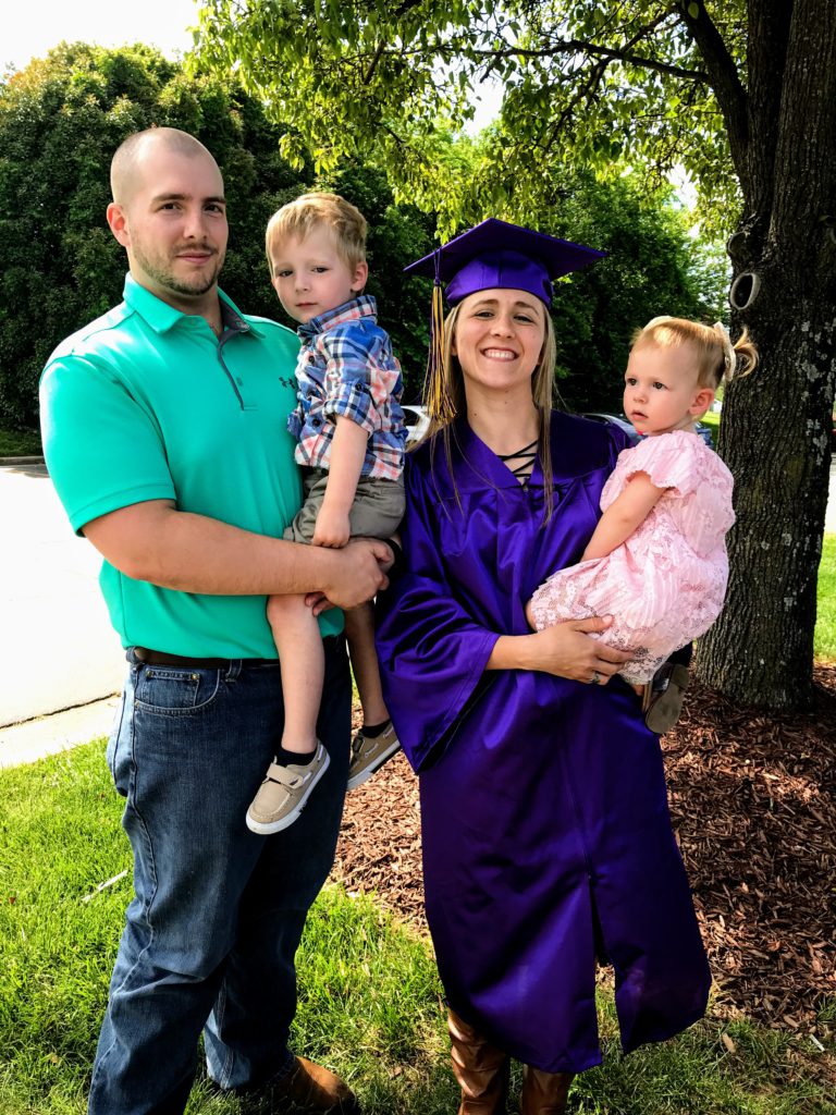 A father and a mother, dressed in a cap and gown, hold their two children